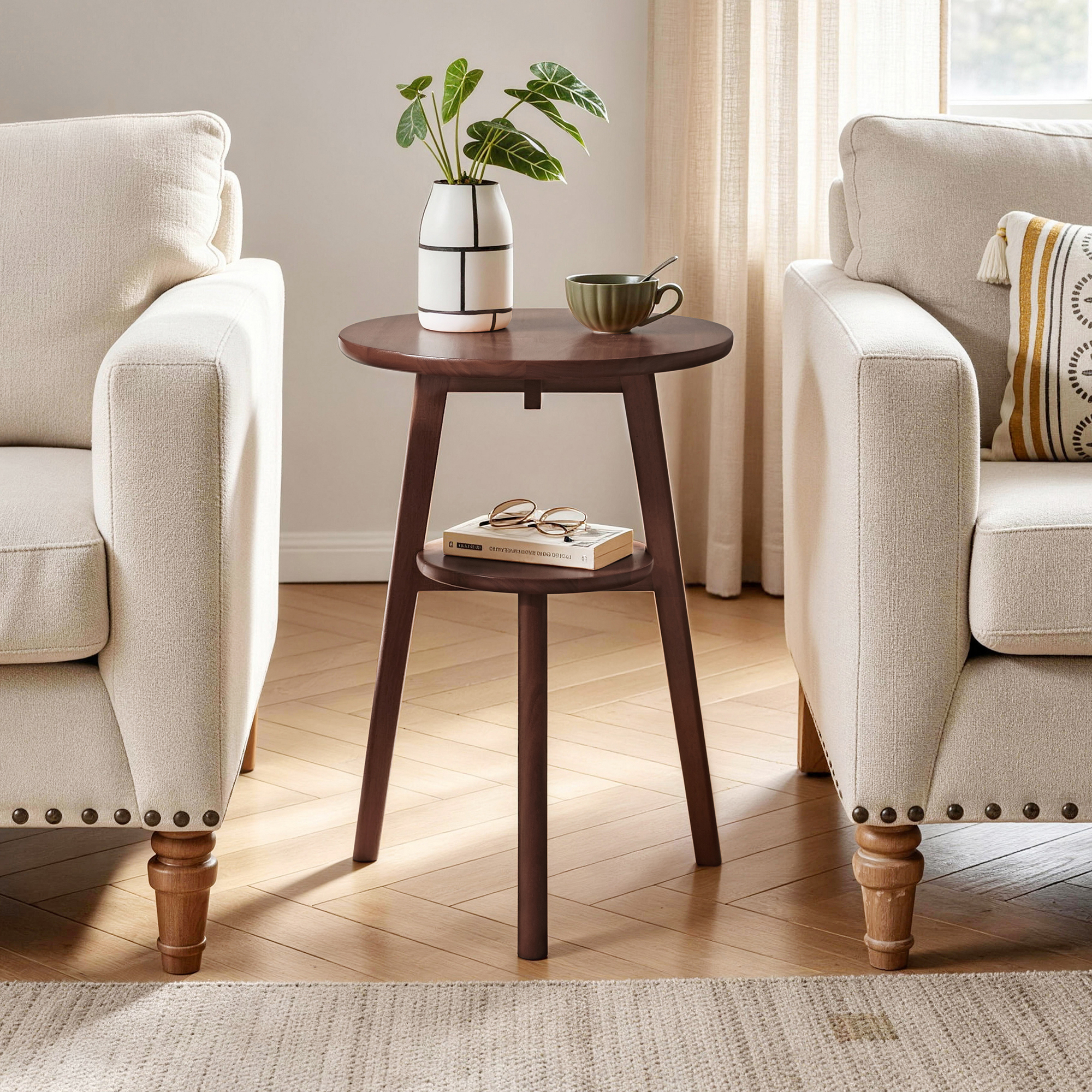 Walnut 2-tier round side table placed between two armchairs with books on the lower shelf