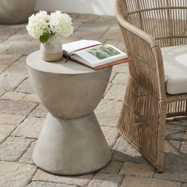 Light grey concrete side table holding a vase of white hydrangeas and a book on a brick patio next to a rattan chair