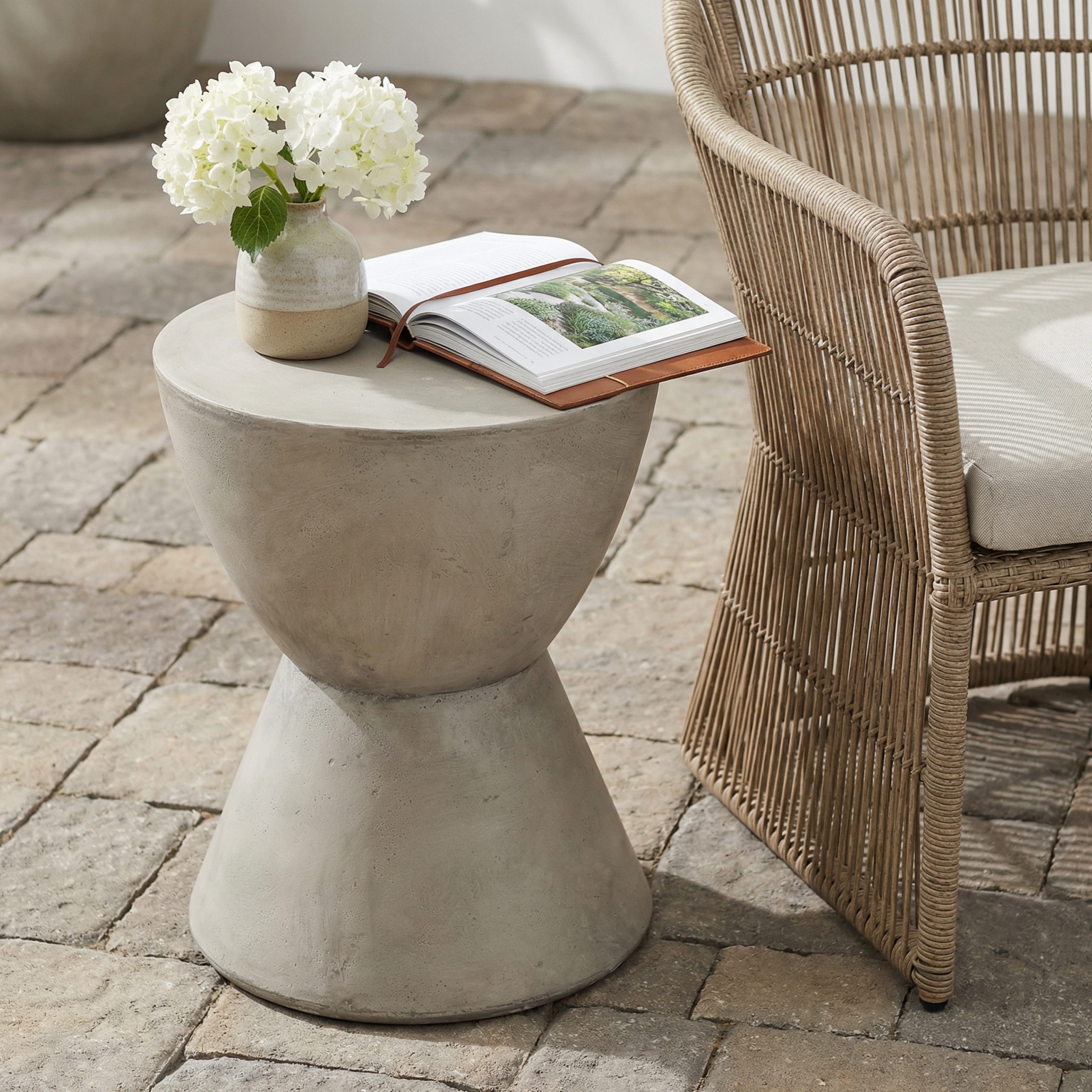 Light grey concrete side table holding a vase of white hydrangeas and a book on a brick patio next to a rattan chair