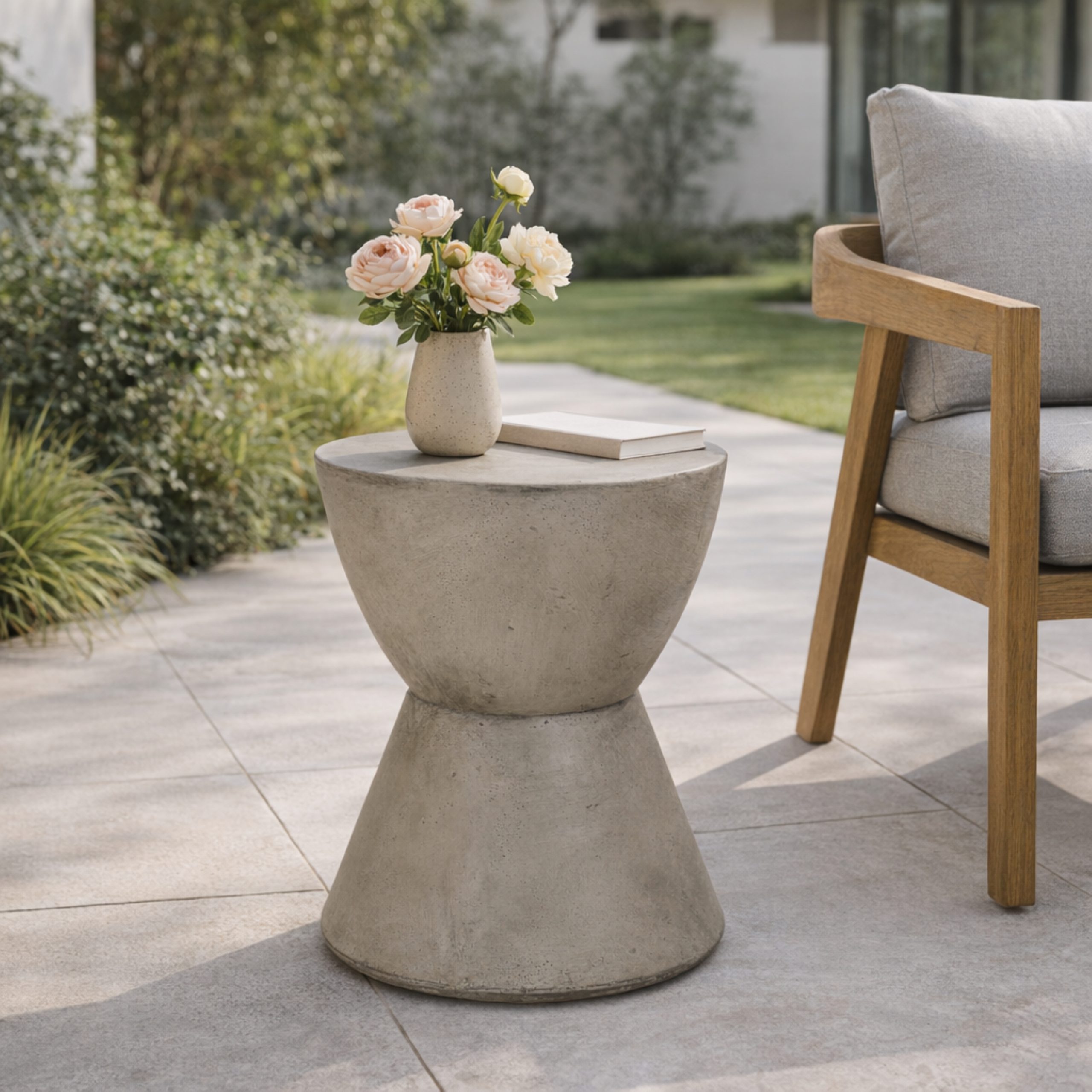 Lightweight concrete patio side table with a small vase of roses beside a teak armchair in a garden