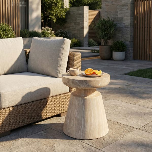 Sculptural pedestal side table in travertine finish holding snacks beside a wicker outdoor sofa on a stone patio