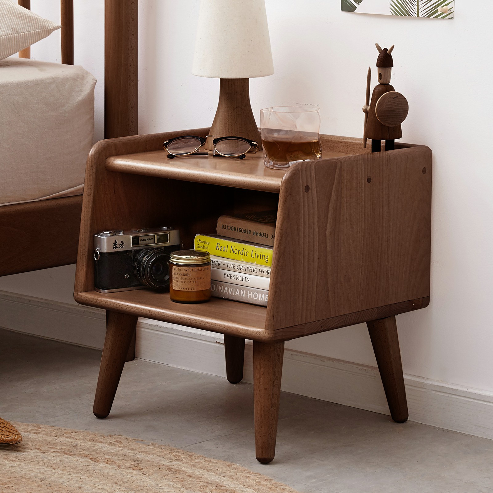 Walnut nightstand with books and camera beside a bed, angled side view